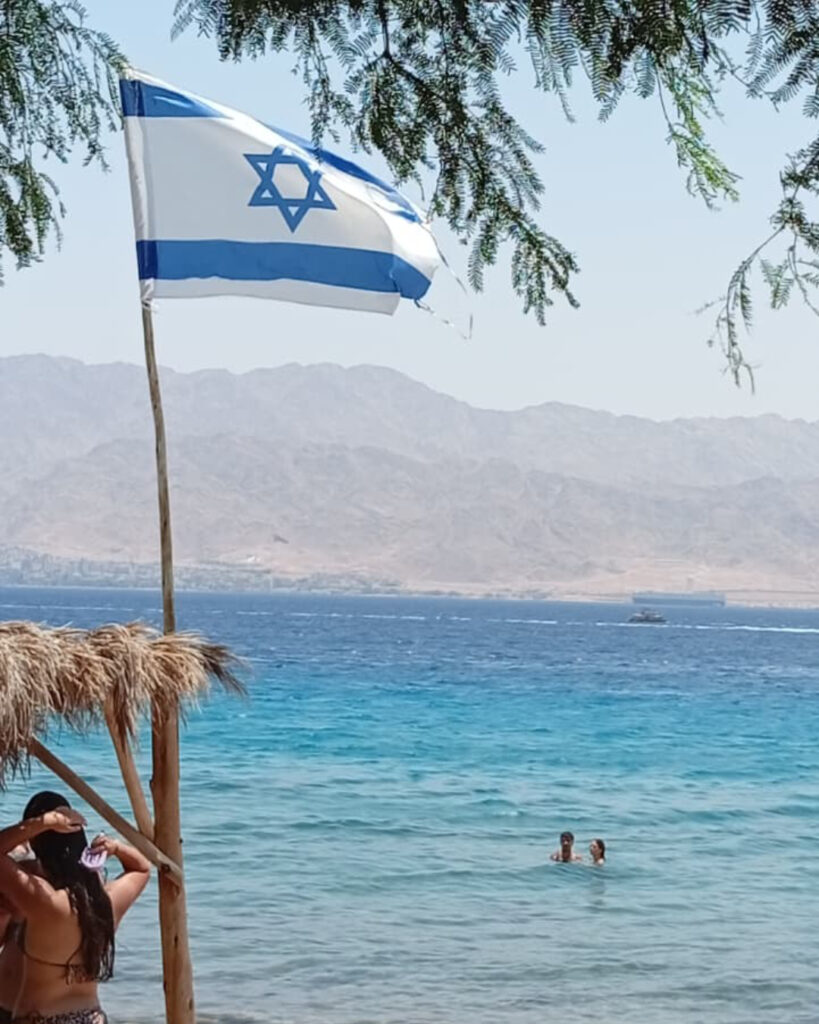 דגל ישראל וזוג במים בחוף של מוש. הרי אדום ברקע